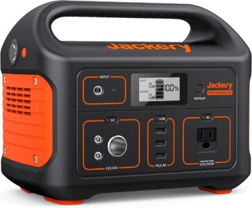 Jackery Portable Power Station Explorer 500 powering a laptop and camping light on a wooden picnic table outdoors.