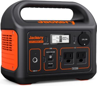 Jackery Explorer 300 Portable Power Station sitting on a forest floor next to a charging drone and a DSLR camera.
